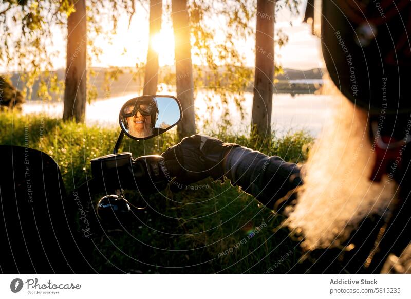 Senior Biker's Reflexion in Motorrad-Spiegel bei Sonnenuntergang Reflexion & Spiegelung Mann Lächeln Schutzhelm Sonnenbrille Vollbart Glück Bäume See Natur
