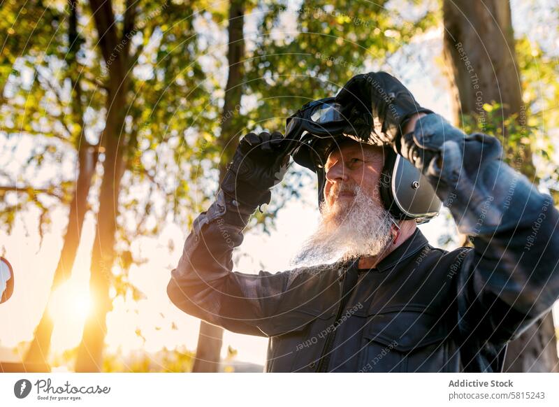 Senior Biker beim Einstellen der Schutzbrille im Freien bei Sonnenuntergang männlich Schutzausrüstung weißer Bart reif Wegsehen Schutzhelm Motorrad Freizeit