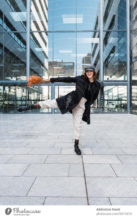 Glückliche Frau mit Blumen tanzt auf der Straße Tanzen Lächeln Blumenstrauß Glaswand Gebäude Stadtzentrum Stil Straßenbelag modern Bein angehoben heiter urban