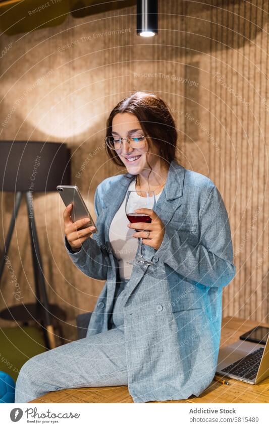 Junge Geschäftsfrau mit Wein und Smartphone benutzend Pause Arbeitsplatz Lächeln Rotwein soziale Netzwerke freiberuflich Tisch Frau jung Laptop online Apparatur