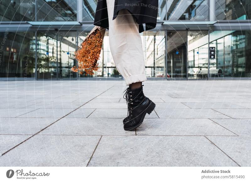 Crop-Frau mit Blumenstrauß auf Zehenspitzen auf der Straße Straßenbelag Stil Gleichgewicht urban Gebäude Stiefel Hose Mantel Bürgersteig Glaswand Zeitgenosse