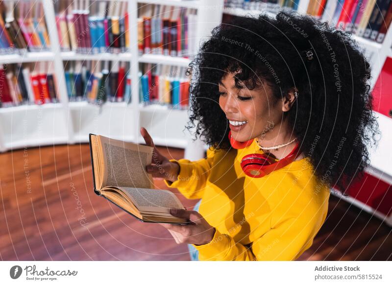 Zufriedene ethnische Studentin beim Lesen eines Lehrbuchs in der Bibliothek Frau lesen Buch Schüler Lächeln Bildung Glück Leseratte Bücherschrank positiv trendy