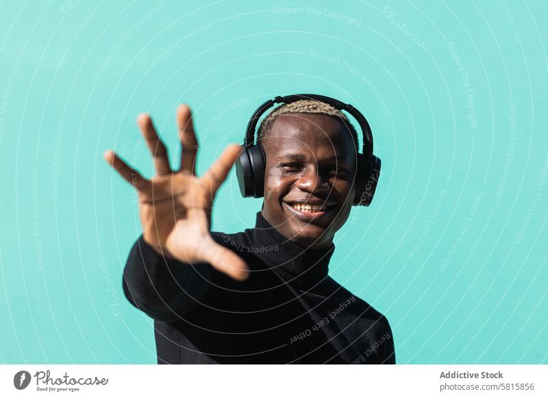 Stilvoller junger schwarzer Mann, der mit Kopfhörern Musik hört und die Hand ausstreckt zuhören gestikulieren Lächeln genießen positiv Porträt Straße Glück