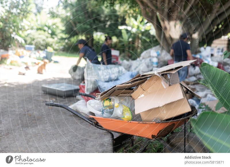 Gepackte Schubkarre mit Kunststoffen und Pappe mit Hintergrundarbeitern verpackt Karton Recycling Arbeitnehmer beladen Aktivitäten Unschärfe engagiert