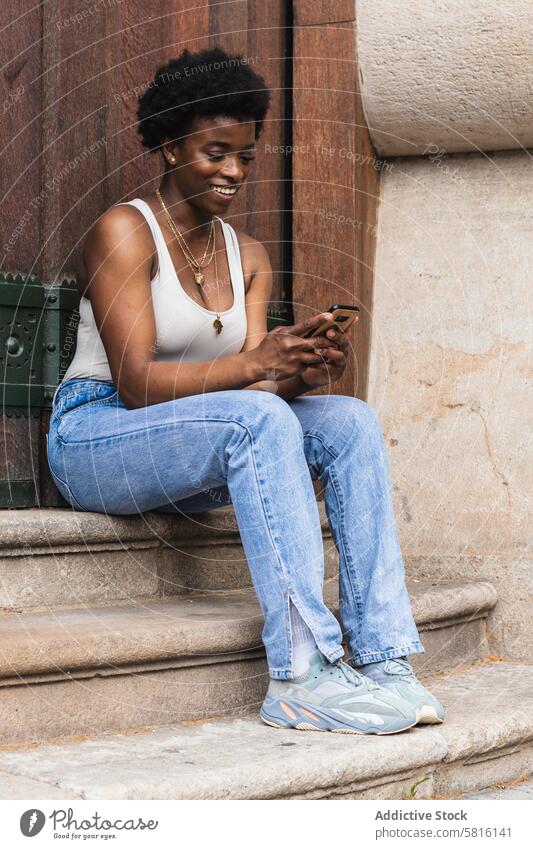 Lächelnde schwarze Frau mit Smartphone sitzt in der Nähe von Gebäude benutzend heiter Surfen Texten sorgenfrei Internet digital Glück soziale Netzwerke genießen