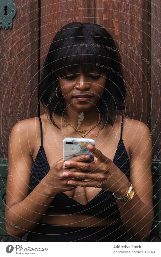 Schwarze Frau, die mit ihrem Smartphone chattet benutzend heiter Dekolleté soziale Netzwerke positiv Spaß Moment Lächeln Gerät Apparatur Glück genießen Stil