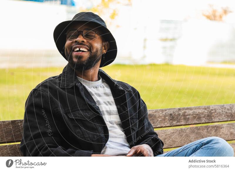 Trendy schwarzer Mann sitzt auf Bank Hipster Glück Park heiter trendy Hut Erwachsener Afroamerikaner ethnisch männlich Lächeln modern online Internet