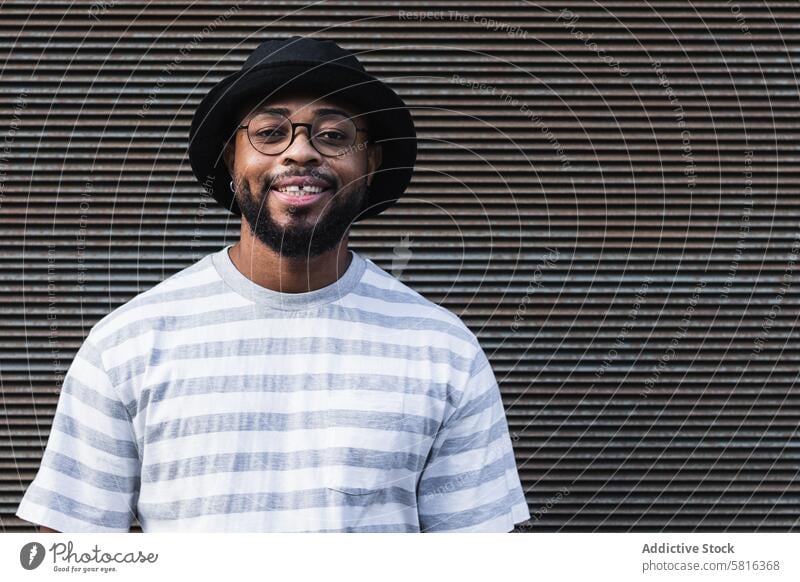 Lächelnder schwarzer Hipster-Mann mit Hut schaut in die Kamera Glück trendy heiter Vollbart Brille Optimist männlich Afroamerikaner ethnisch Outfit Stil positiv