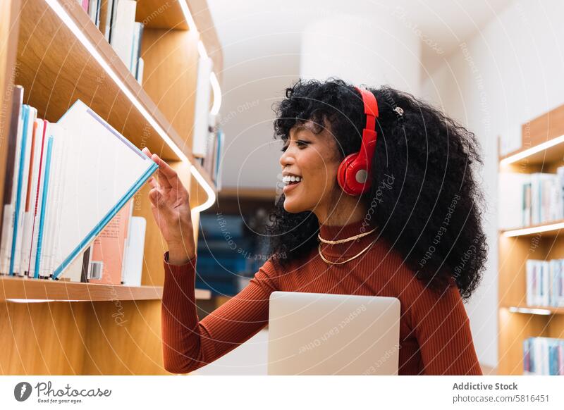 Zufriedene Frau mit Kopfhörern, die ein Buch in der Bibliothek liest melomaniac Regal Schüler Bücherschrank positiv wählen Bücherregal Lächeln Musik zuhören