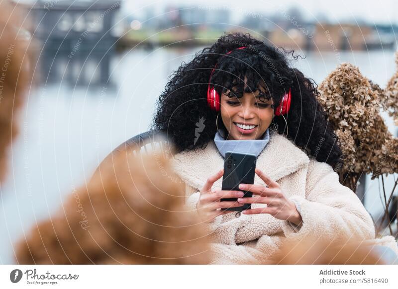 Fröhliche schwarze Frau, die am Seeufer Musik hört Kopfhörer Smartphone benutzend zuhören Kälte ruhen sich[Akk] entspannen Park afroamerikanische Frau Großstadt