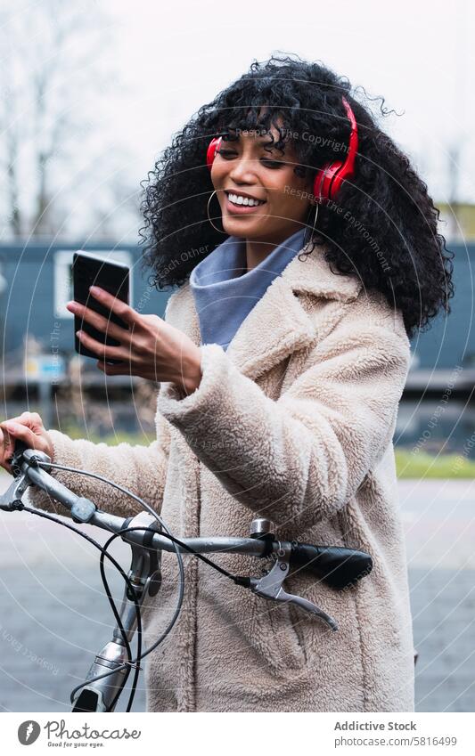 Vergnügte schwarze Frau mit Smartphone auf dem Fahrrad Kopfhörer benutzend Musik zuhören Mitfahrgelegenheit Kälte Spaziergang afroamerikanische Frau