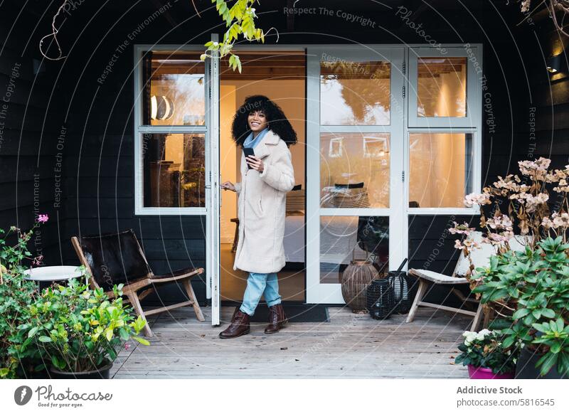 Glückliche schwarze Frau steht in der Tür eines Hauses heimwärts hinausgehend warme Kleidung Mantel Oberbekleidung Türöffnung urban Smartphone Telefon