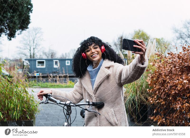 Schwarze Frau mit Smartphone auf dem Fahrrad unter Selfie Spaziergang Kopfhörer benutzend Musik zuhören Mitfahrgelegenheit Kälte afroamerikanische Frau