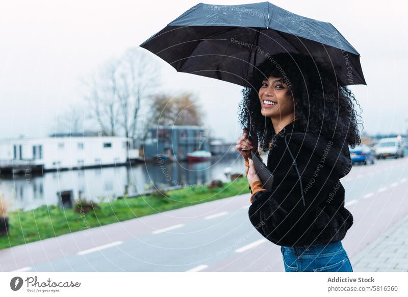 Lächelnde schwarze Frau mit Regenschirm auf der Straße Großstadt Straßenrand urban Vorschein Tablette Porträt afroamerikanische Frau lässig jung Glück positiv