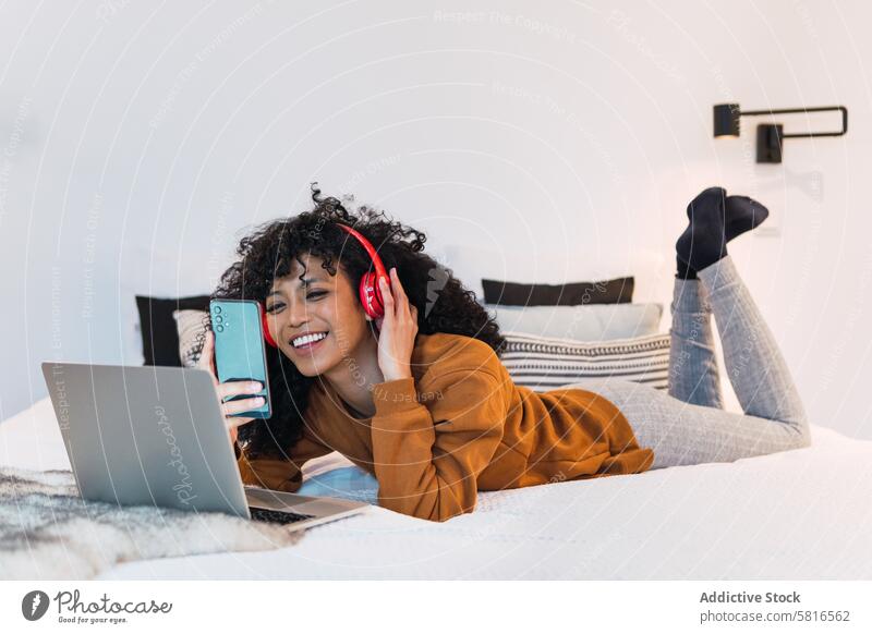 Fröhliche schwarze Frau mit Laptop und Smartphone Kopfhörer benutzend Musik Netbook ruhen sich[Akk] entspannen Kälte Porträt Afroamerikaner Bett Telefon