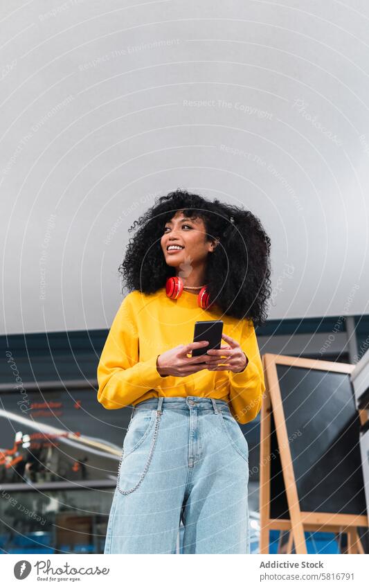 Fröhliche, stilvolle ethnische Dame, die in einem modernen Gebäude steht und ein Smartphone benutzt Frau Lächeln stehen Kommunizieren selbstbewusst trendy