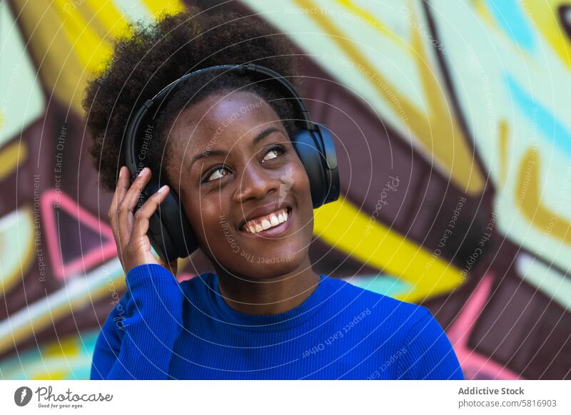 Positive ethnische Frau genießt Musik in der Stadt Kopfhörer zuhören Graffiti Wand Großstadt cool tausendjährig urban schwarz Afroamerikaner Afro-Look Frisur