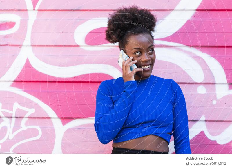 Zufriedene schwarze Frau spricht in der Stadt über ihr Smartphone sprechen Graffiti Wand Großstadt Straßenkunst heiter diskutieren reden ethnisch Afroamerikaner