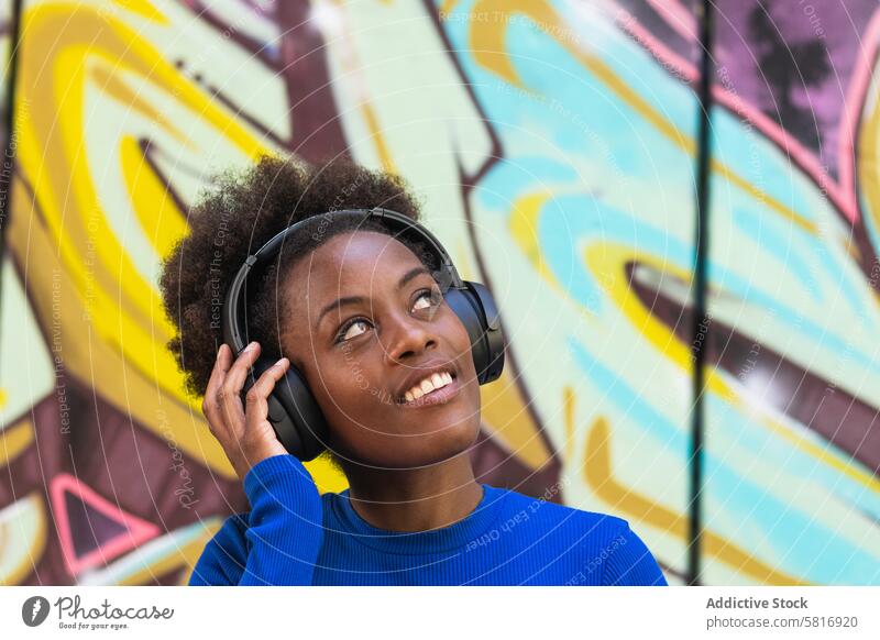 Positive ethnische Frau genießt Musik in der Stadt Kopfhörer zuhören Graffiti Wand Großstadt cool tausendjährig urban schwarz Afroamerikaner Afro-Look Frisur