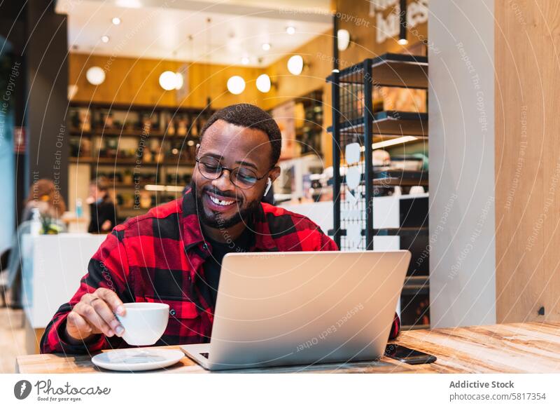 Lächelnder schwarzer Mann arbeitet an einem Laptop in einem Café Kopfhörer benutzend online Internet Browsen freiberuflich Arbeit männlich Afroamerikaner