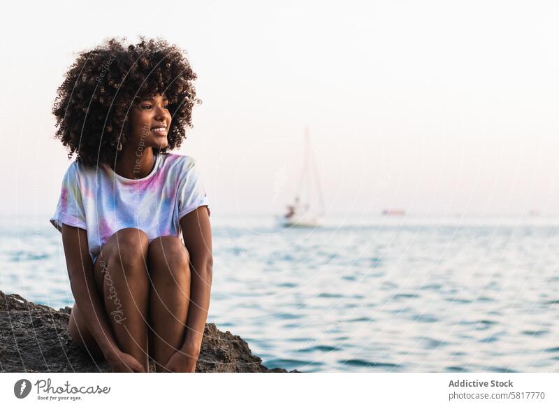 Entzückte ethnische Frau lacht am Meer Strand Lachen Sommer Meeresufer MEER schlank Afro-Look Frisur Spaß haben schwarz Afroamerikaner Felsen sitzen T-Shirt