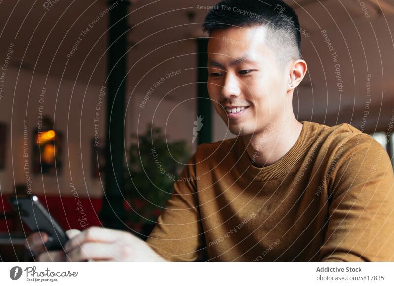 Lächelnder asiatischer Mann sitzt im Restaurant und schaut auf sein Smartphone. jung Lifestyle Glück Kaffee Menschen Café Sitzen Technik & Technologie Telefon