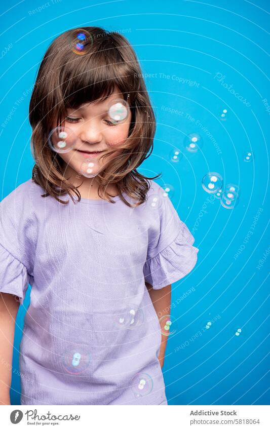 Fröhliches Mädchen spielt mit Seifenblasen vor blauem Hintergrund Kind Schaumblase spielen Spaß Freude heiter Studioaufnahme Blauer Hintergrund purpur Hemd