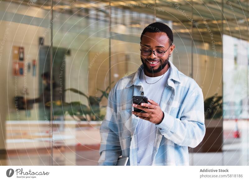 Fröhlicher schwarzer Mann, der in der Nähe eines Gebäudes auf seinem Smartphone surft Browsen Textnachricht online Talkrunde Internet Straße Glaswand Großstadt