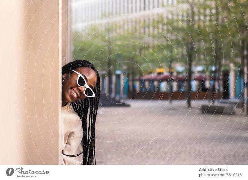 Fröhliche ethnische Frau lugt hinter einer Säule hervor schauen Sie Spalte Straße Lächeln Großstadt Stil Spaß Gebäude jung Glück schwarz Afroamerikaner heiter