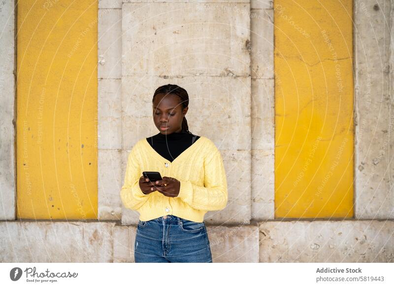 Junge Frau im gelben Pullover schreibt eine SMS auf dem Smartphone Texten Beton Wand Jeanshose jung pulsierend Fokus im Freien Technik & Technologie Mitteilung