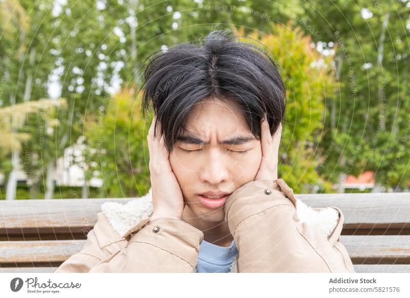 Junger Mann mit Kopfschmerzsymptomen im Freien Kopfschmerzen Unbehagen Hypoglykämie Symptome jung asiatisch Park Bank umklammert Schmerz Gesundheit medizinisch