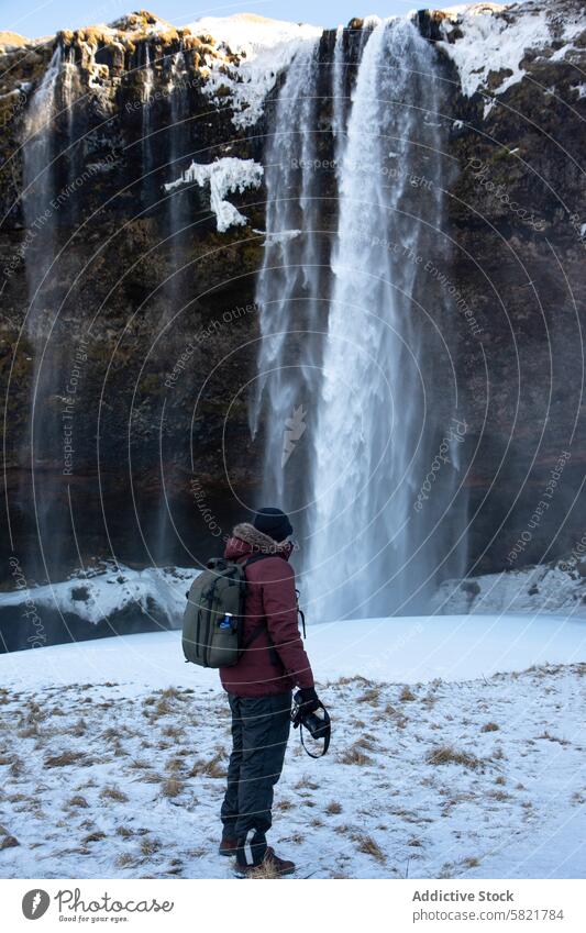 Mann mit Kamera vor majestätischem Wasserfall in verschneitem Island Fotograf Fotokamera Rucksack Natur im Freien Winter Schnee Eis reisen Erkundung malerisch