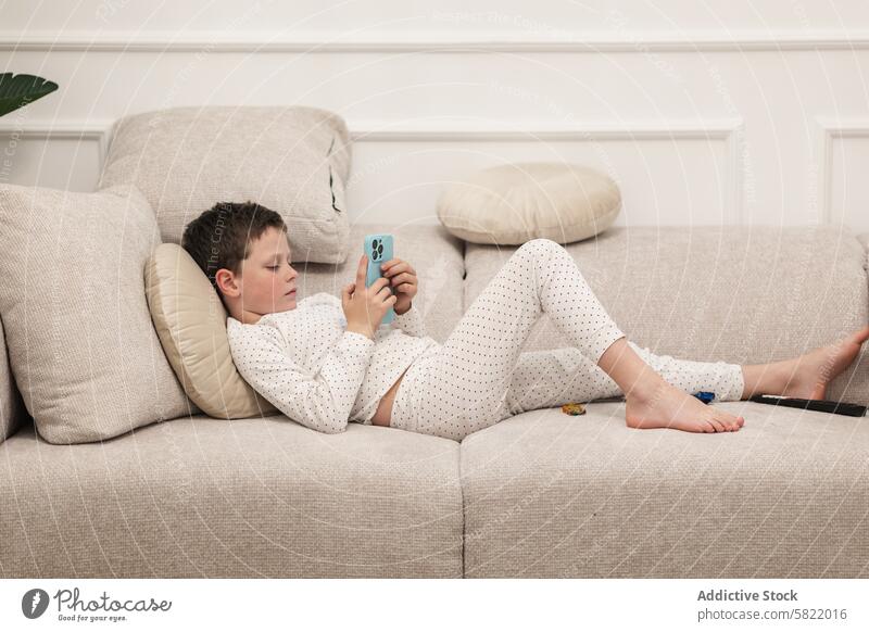 Junge im Schlafanzug mit Smartphone auf der Couch zu Hause Kind heimwärts Pyjama Punktmuster Liege räkeln fokussiert im Innenbereich legt sich hin
