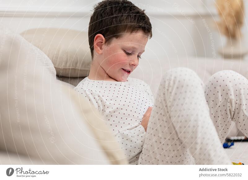 Junge im Schlafanzug genießt seine Freizeit zu Hause Kind heimwärts Pyjama Sitzen Liege Gerät Handheld Spielen Wegsehen bequem Freude im Innenbereich Youngster