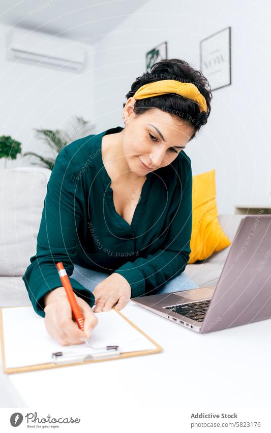 Frau arbeitet von zu Hause aus mit Laptop und Klemmbrett Zwischenablage heimwärts Arbeit schreibend Hinweis sitzend grüne Bluse heller Raum