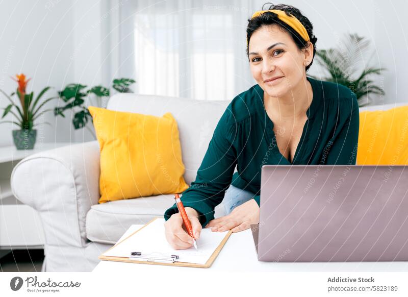 Frau schreibt auf einem Klemmbrett, das neben einem Laptop zu Hause steht schreibend Zwischenablage heimwärts Lächeln selbstbewusst grüne Bluse Notizen