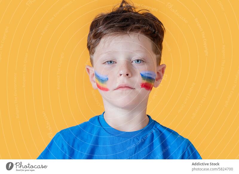 Junge mit buntem Gesicht malen auf einem gelben Hintergrund Gesichtsfarbe gelber Hintergrund Porträt farbenfroh Kind ernst blaues Hemd Farbe jung feierlich