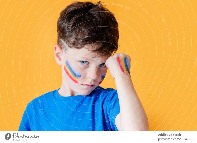 Junger Junge mit Regenbogenfarbe im Gesicht vor gelbem Hintergrund Kind Gesichtsfarbe Ausdruck Vielfalt Jugend Farbe pulsierend gelber Hintergrund Augenkontakt