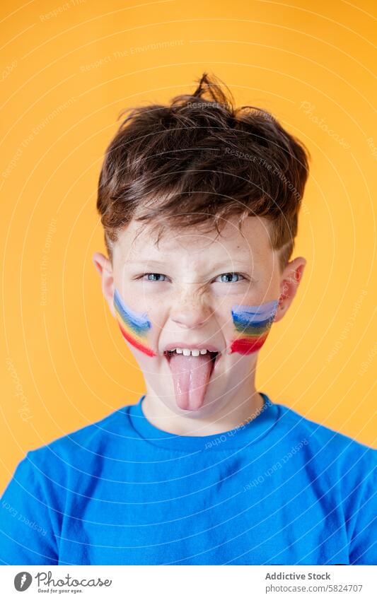 Junge mit bunter Gesichtsbemalung streckt die Zunge heraus Gesichtsfarbe spielerisch oranger Hintergrund blaues Hemd Kind Regenbogen farbenfroh Wange Farbe hell