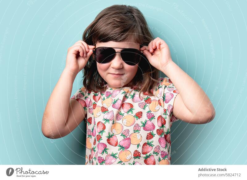 Junges Mädchen mit Sonnenbrille posiert mit einem zuversichtlichen Lächeln jung Kind Mode selbstbewusst blau Hintergrund heiter Stil farbenfroh Frucht Muster