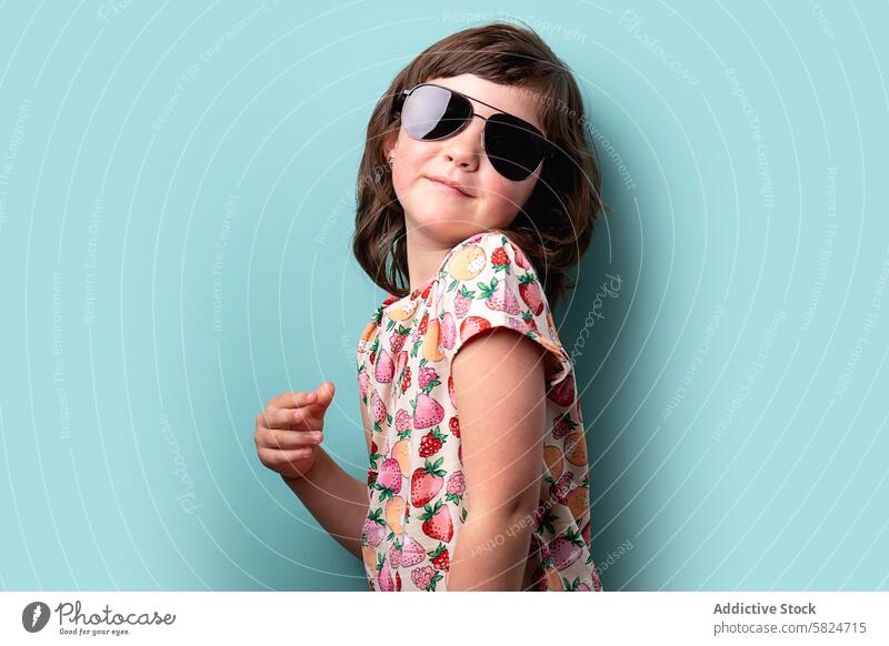 Junges Mädchen mit Sonnenbrille und gemustertem Kleid posiert spielerisch Kind Pose Lächeln selbstbewusst heiter farbenfroh Blauer Hintergrund Muster Frucht