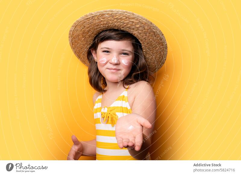 Junges Mädchen mit Sonnenhut und Sonnenschutzmittel auf gelbem Hintergrund Strohhut pulsierend heiter Badeanzug gestreift posierend Sommer Schutz Hautpflege