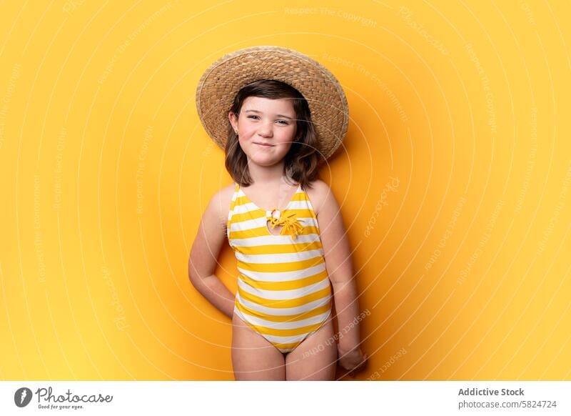 Junges Mädchen mit Strohhut und gestreifter Badehose auf gelbem Hintergrund Badeanzug Sommer heiter Pose Kind Unschuld pulsierend Textur Mode Freizeit