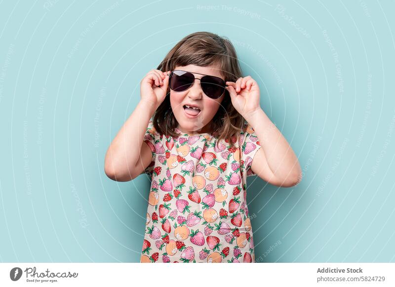 Junges Mädchen lächelt und rückt ihre Sonnenbrille zurecht Kind Lächeln Mode spielerisch Einstellung blaugrün Hintergrund jung freudig Frucht Muster Sommer Stil
