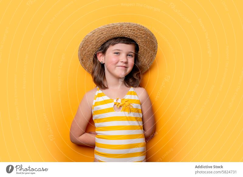 Lächelndes junges Mädchen im gestreiften Badeanzug und Strohhut gelber Hintergrund Kind heiter Fröhlichkeit Sommer Porträt Mode Jugend lässig niedlich Unschuld