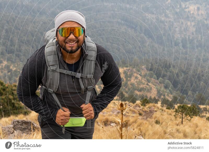 Lächelnder Wanderer mit Rucksacktraining im Gebirge Berge u. Gebirge Wanderung Training männlich Beanie Sonnenbrille heiter Abenteuer Gelände Fitness Reise