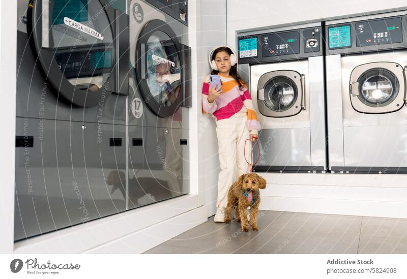 Preteen-Mädchen mit Hund wartet in einem Waschsalon Kind Kopfhörer Smartphone Warten anleinen Haustier Waschmaschine im Innenbereich lässig Kleidung Wäscherei