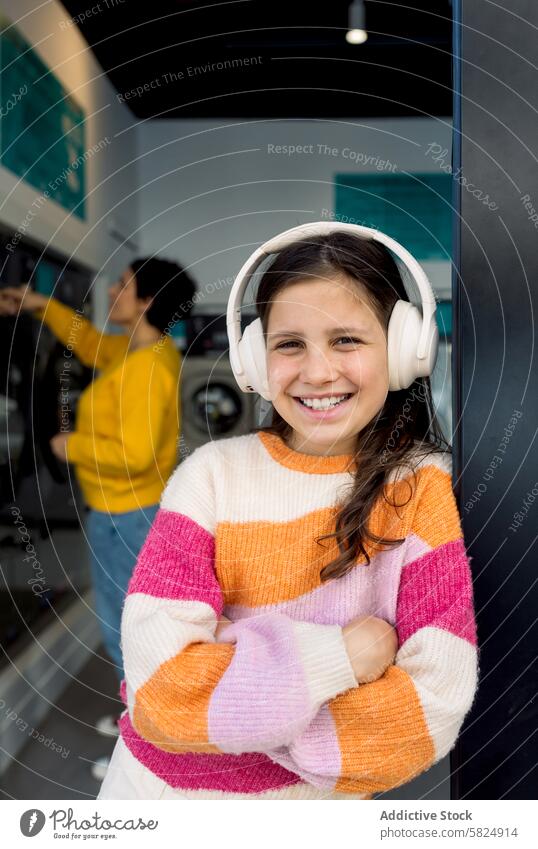 Preteen-Mädchen genießt Musik in der Wäscherei mit lächelnden Erwachsenen Kind Kopfhörer Lächeln Erwachsener Kleidung Sortierung Lehnen Maschine Hintergrund