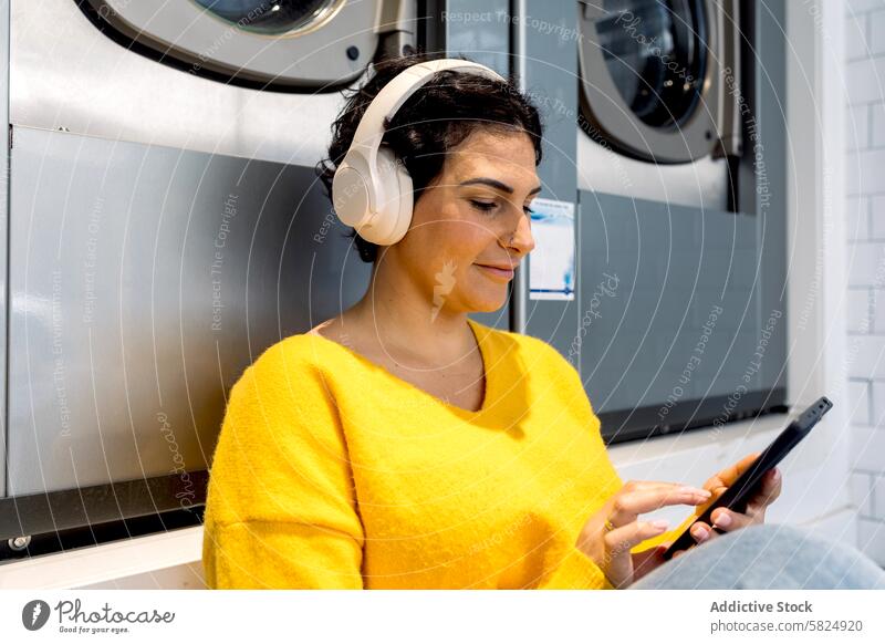 Frau entspannt sich bei Musik im Waschsalon Erwachsener Kopfhörer Sitzen Smartphone Wäscherei Waschmaschine Handy Freizeit Technik & Technologie Erholung lässig