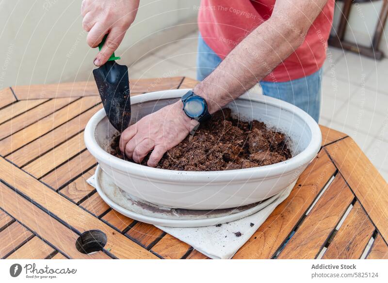 Mann pflanzt in einem weißen Behälter auf einem Holztisch Bepflanzung Container hölzern Tisch Kelle Boden im Freien Gartenarbeit Hobby Natur Armbanduhr lässig